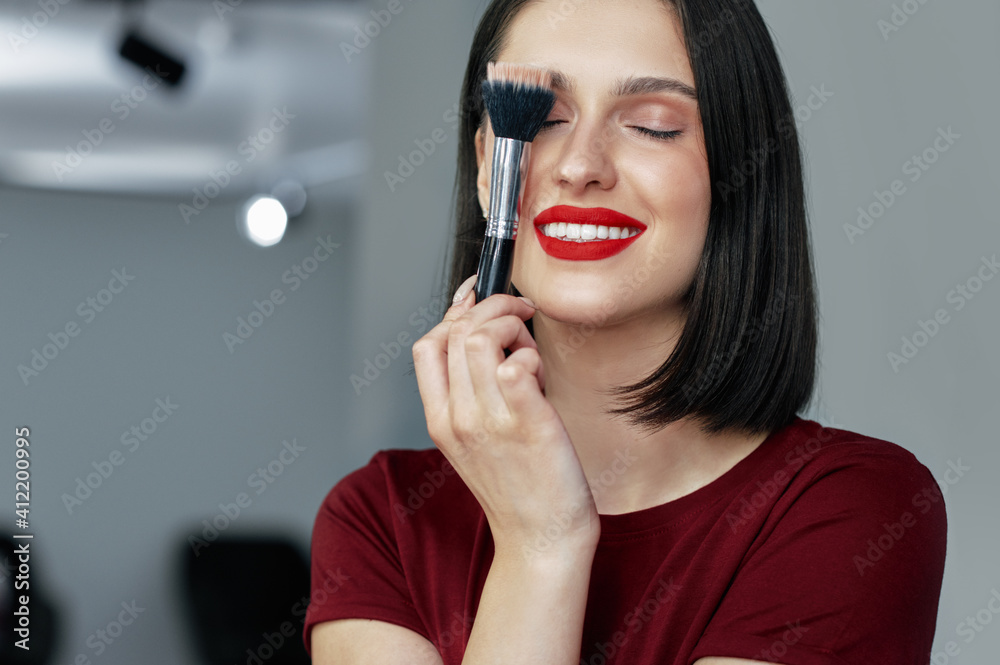 Candid portrait of a gorgeous brunette female with red lips posing with a brush after making make-up tutorial. Happy young woman making herself makeup with a brush with organic cosmetics. Candid portrait of a gorgeous brunette female with red lips posing with a brush after making make-up tutorial. Happy young woman making herself makeup with a brush with organic cosmetics.