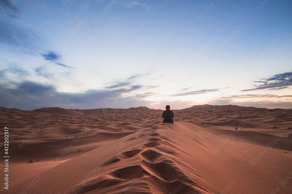 A young female traveller enjoys a sunrise or sunset landscape view of the desert sand dunes of Erg Chebbi near the village of Merzouga, Morocco.