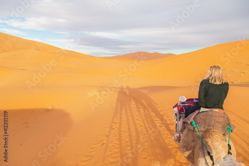 A young blonde female tourist on a camelback excursion along the desert sand dunes of Erg Chebbi near the village of Merzouga in southeastern Morocco.