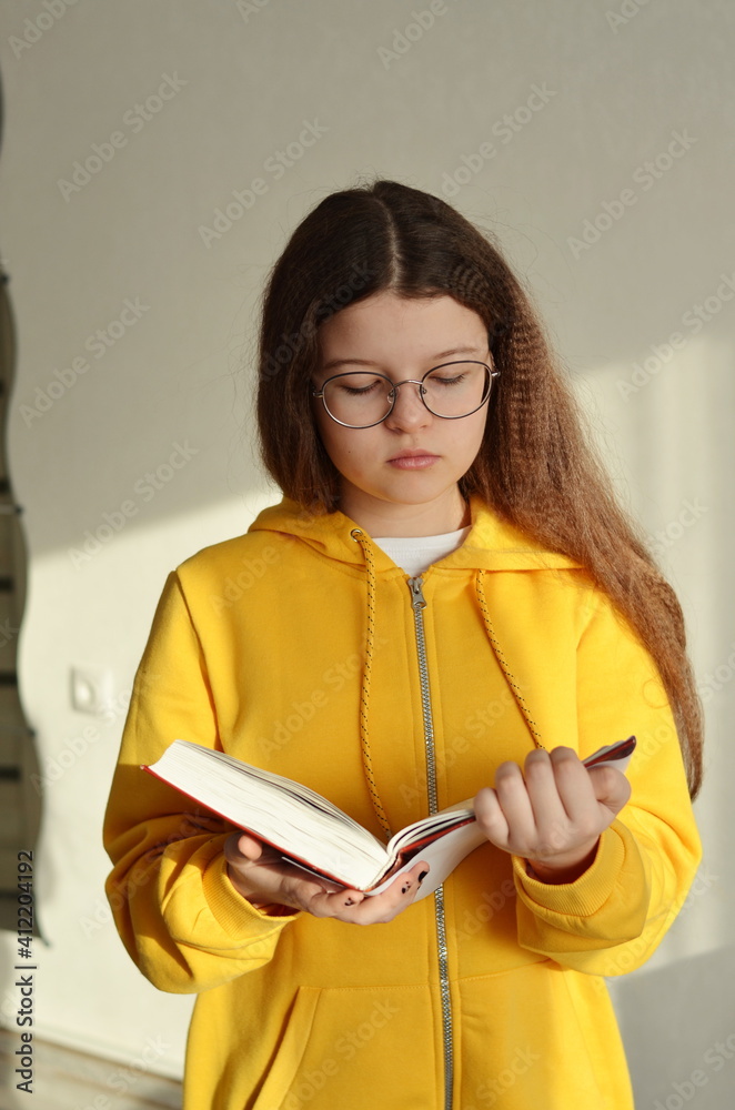 Beautiful girl with glasses reading a book.Shes wearing a yellow hoodie.The girl has long brown hair. Beautiful girl with glasses reading a book.Shes wearing a yellow hoodie.The girl has long brown hair.