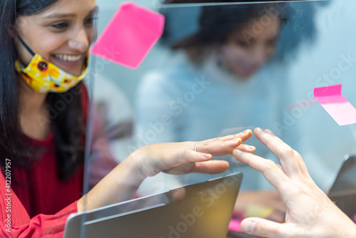 young indian woman  wearing facemask below chin  trying to touch the hand of her colleague in front of her  plexiglas barrier to avoiding direct contact and preventing spreading of Corona Virus