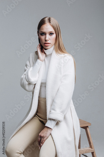 young fashionable beautiful girl in a trendy white coat posing on a gray background