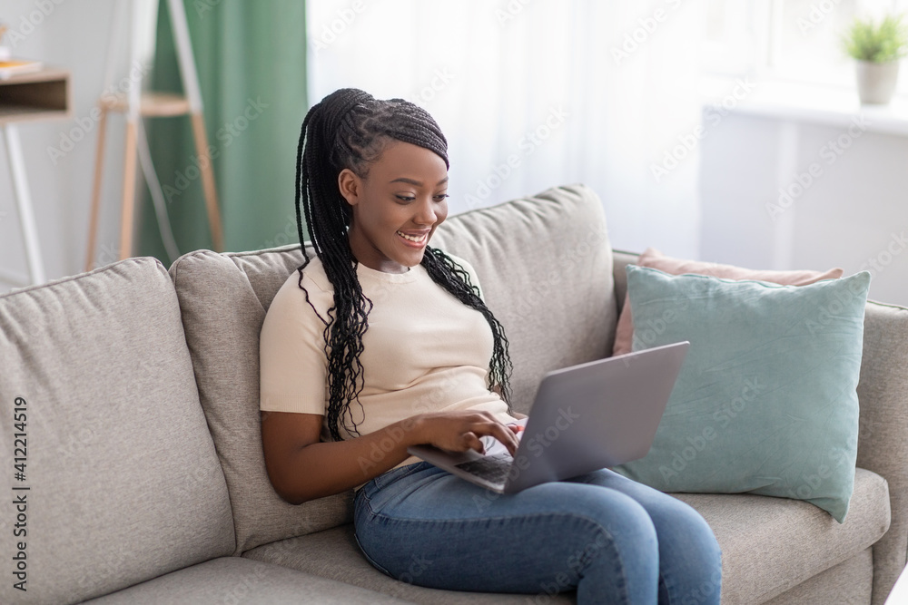 Obraz premium Joyful african lady sitting on couch at home, using laptop