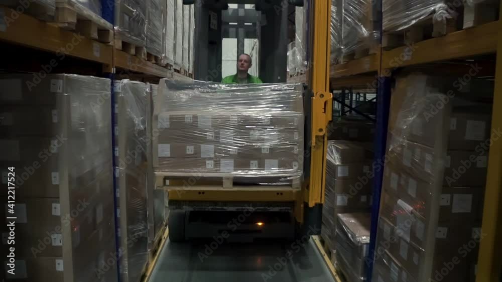 Man worker lifting cargo using loading equipment during working day at warehouse. Front view of young male employee is moving while sitting in car, doing work in modern storehouse. Concept: business Man worker lifting cargo using loading equipment during working day at warehouse. Front view of young male employee is moving while sitting in car, doing work in modern storehouse. Concept: business