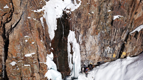 Freezing waterfall in the snowy mountains. View from the drone, from above. The rocks are covered with snow and ice. A small stream of water runs. The waterfall freezes. A group of people are resting photo