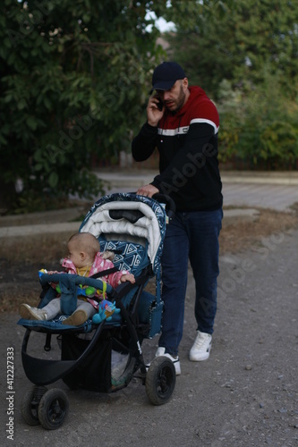 a young dad pushes a stroller and a baby and talks on the phone