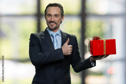 Senior manager with red gift box and showing thumb up. Standing indoors, blurred windows on the background. photo