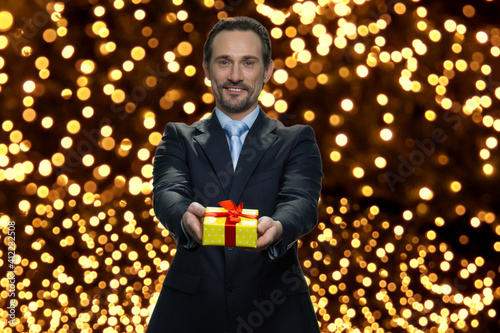 Businessman is giving yellow gift box with red ribbon. Confident senior manager is congratulating you with christmas. Many lights on the background. photo