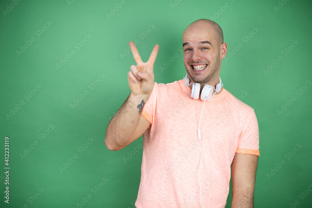 Naklejka premium Young bald man using headphones over green background showing and pointing up with fingers number two while smiling confident and happy