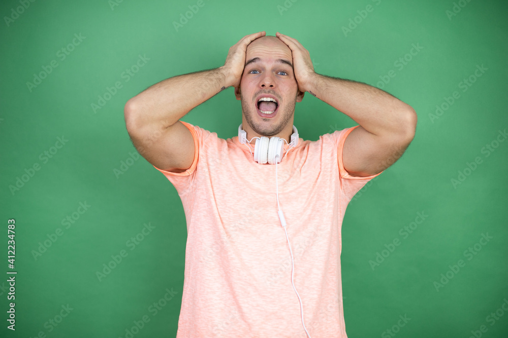Young bald man using headphones over green background crazy and scared with hands on head, afraid and surprised of shock Young bald man using headphones over green background crazy and scared with hands on head, afraid and surprised of shock