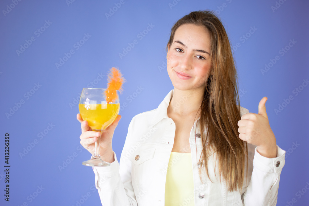 Portrait of a cute casual girl drinking orange juice from a glass and looking at camera isolated over lilac background Portrait of a cute casual girl drinking orange juice from a glass and looking at camera isolated over lilac background