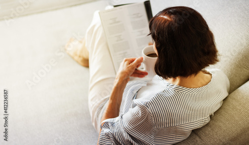 Middle-aged brunette woman with glasses on the gray sofa reading magazine with cup of coffee, soft focus