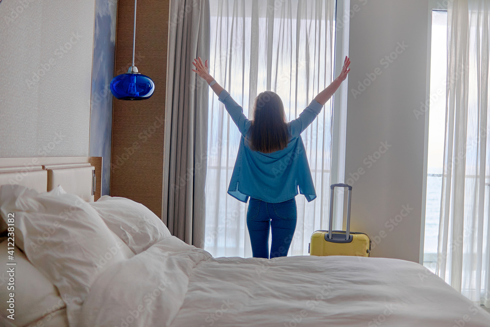 Happy traveler woman with raised hands and suitcase standing near window and looking to a view from hotel room after check-in. Conceptual of travel and vacation holiday - obrazy, fototapety, plakaty 