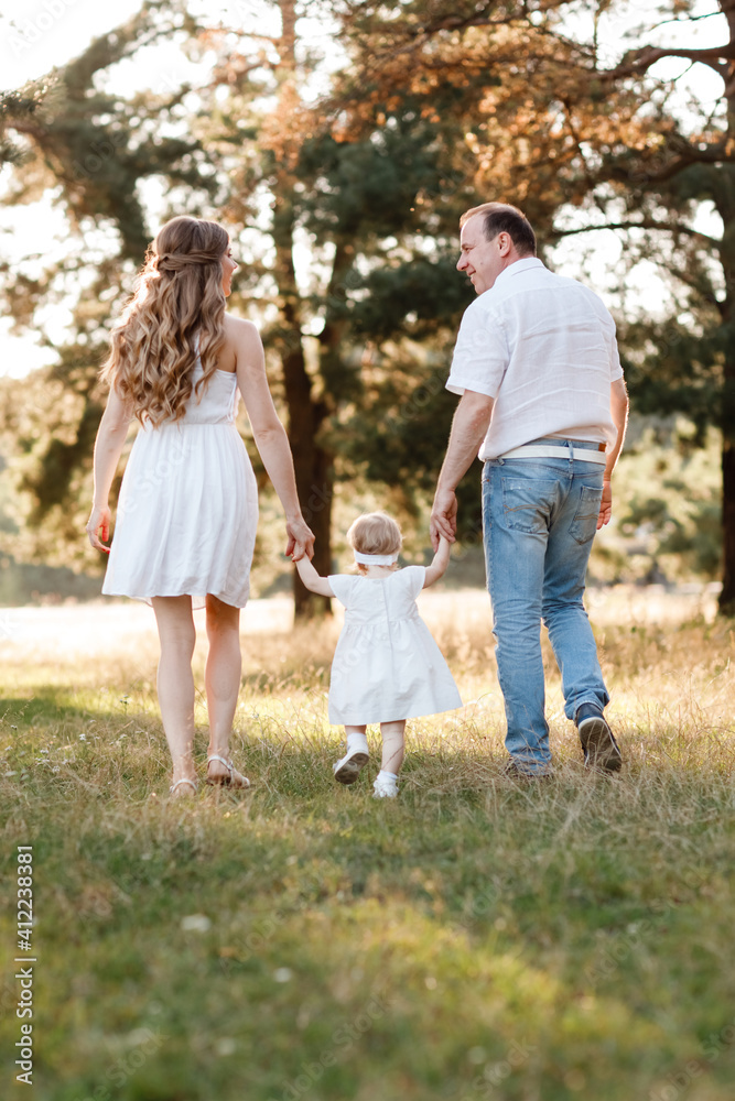 Back view of mother, father hold hands daughter enjoy nature and walk in the summer park. Young family spending time together on vacation, outdoors. Mother's, father's, baby's day Back view of mother, father hold hands daughter enjoy nature and walk in the summer park. Young family spending time together on vacation, outdoors. Mother's, father's, baby's day