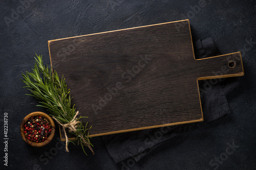 Food cooking background with wooden cutting board, herbs and spices on black stone table. Top view with copy space.