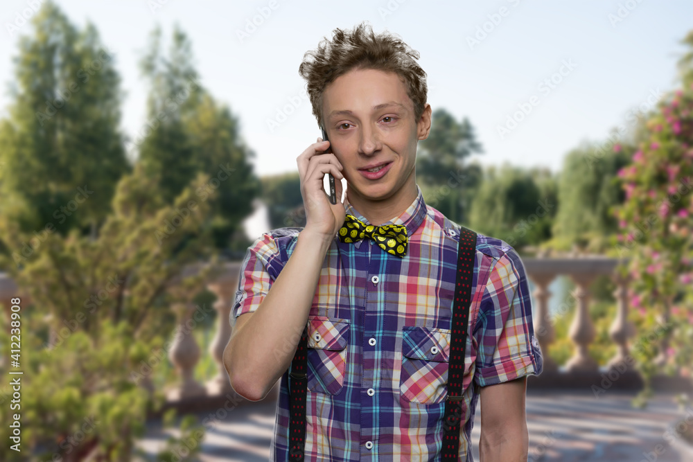 Smiling boy is is talking on phone. Well-dressed young guy has a conversation. Beautifyul summer's nature on the background.