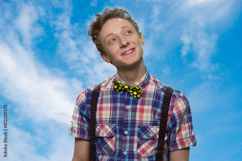 Smiling young boy is dreaming. Thoughtful teen with bowtie standing against blue sky background.