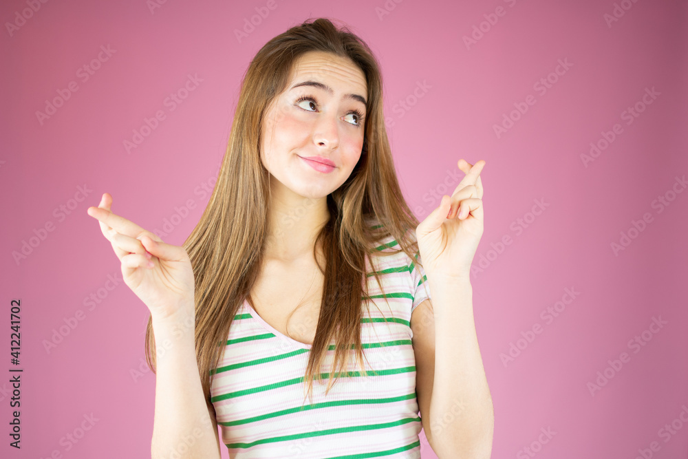 Portrait of a smiling young girl with long hair standing over pink background, holding fingers crossed for good luck