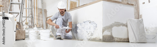 plasterer man at work, take the mortar from the bucket with trowel to plastering the wall of interior construction house site and wear helmet, panoramic image photo