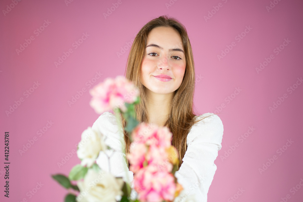 Portrait of a beautiful young girl in white shirt holding big bouquet of flowers isolated over pink background