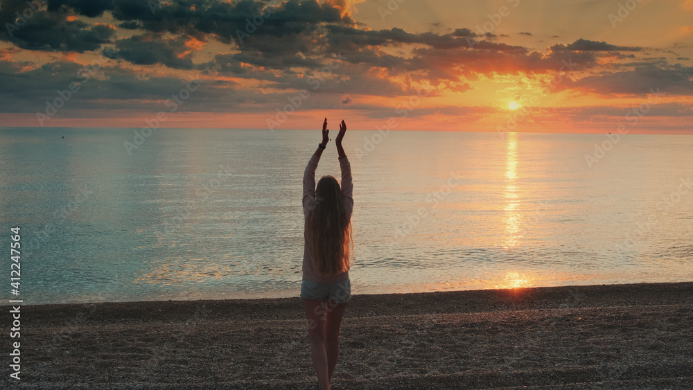 Woman with long hair walking to the sea and raising her hands up. Looking on sunset. Back view.