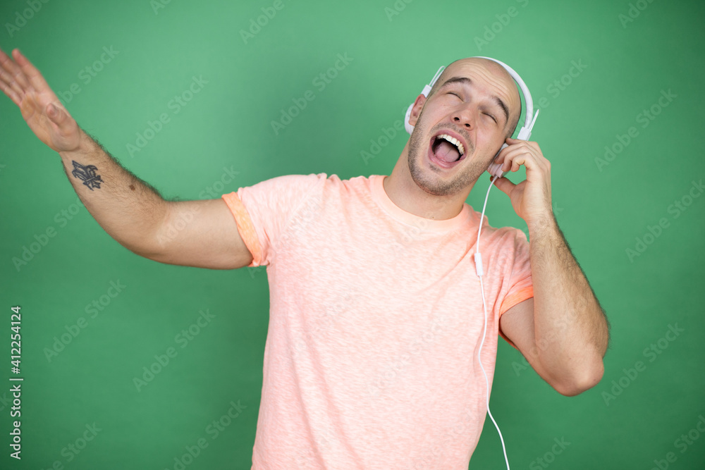 Young bald man using headphones over green background listening to music