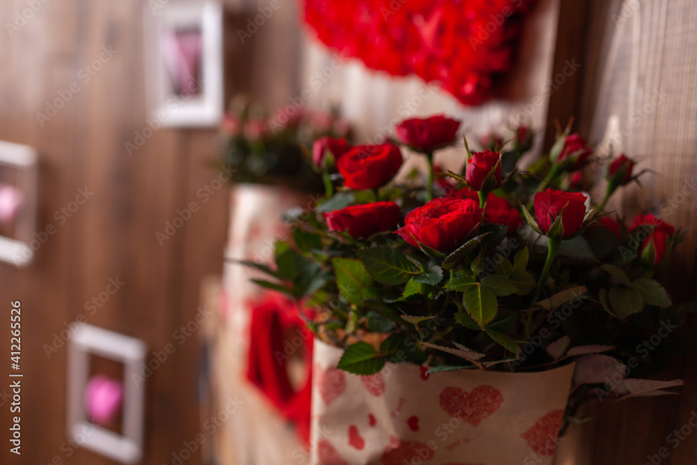 Red and pink potted roses stand on a wooden shelf against a background of brown wooden walls. A potted plant in a festive package. Gifts for Valentine's Day-a red rose in a pot.