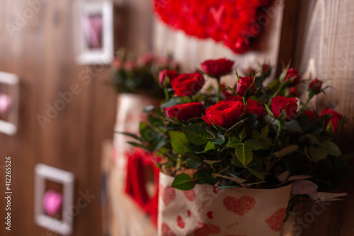 Fototapeta Naklejka Na Ścianę i Meble -  Red and pink potted roses stand on a wooden shelf against a background of brown wooden walls. A potted plant in a festive package. Gifts for Valentine's Day-a red rose in a pot.