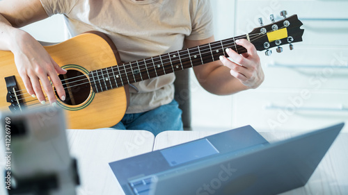 A woman sits in the kitchen during a remote acoustic guitar lesson. A girl learns to play the guitar and watches educational videos on a laptop