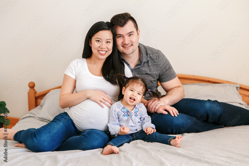 Family Asian Chinese pregnant woman and Caucasian man with mixed race toddler girl sitting on bed at home. Mother, father, baby daughter expecting a new family member. Ethnic diversity.