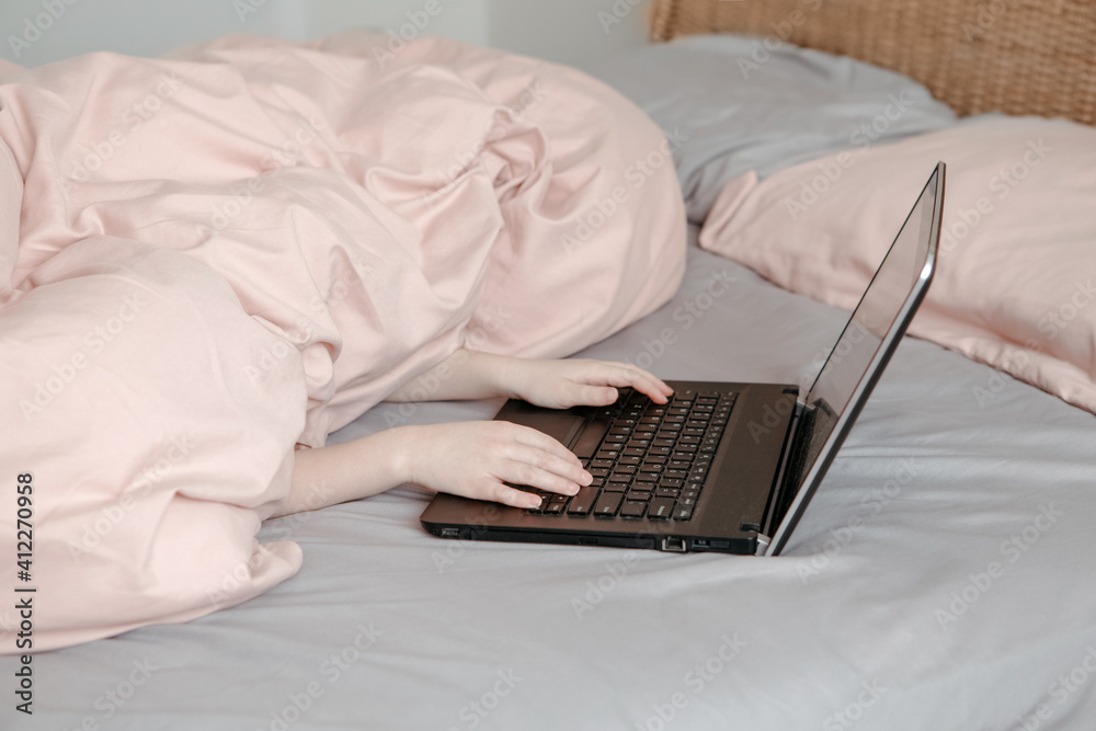 Bored funny child lying in bed under blanket and learning in virtual online school class. Hands typing on computer keyboard. Kid working on a laptop Internet at home. Homeschooling for children. Bored funny child lying in bed under blanket and learning in virtual online school class. Hands typing on computer keyboard. Kid working on a laptop Internet at home. Homeschooling for children.