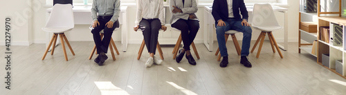 People are waiting in the waiting room. Cropped image of the legs of various people sitting on chairs and waiting their turn for an interview. Concept of employment, clients and human resources. © Studio Romantic