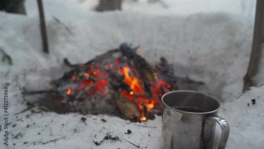 A metal mug with hot tea in front of a campfire in a winter forest. A metal mug with hot tea in front of a campfire in a winter forest.
