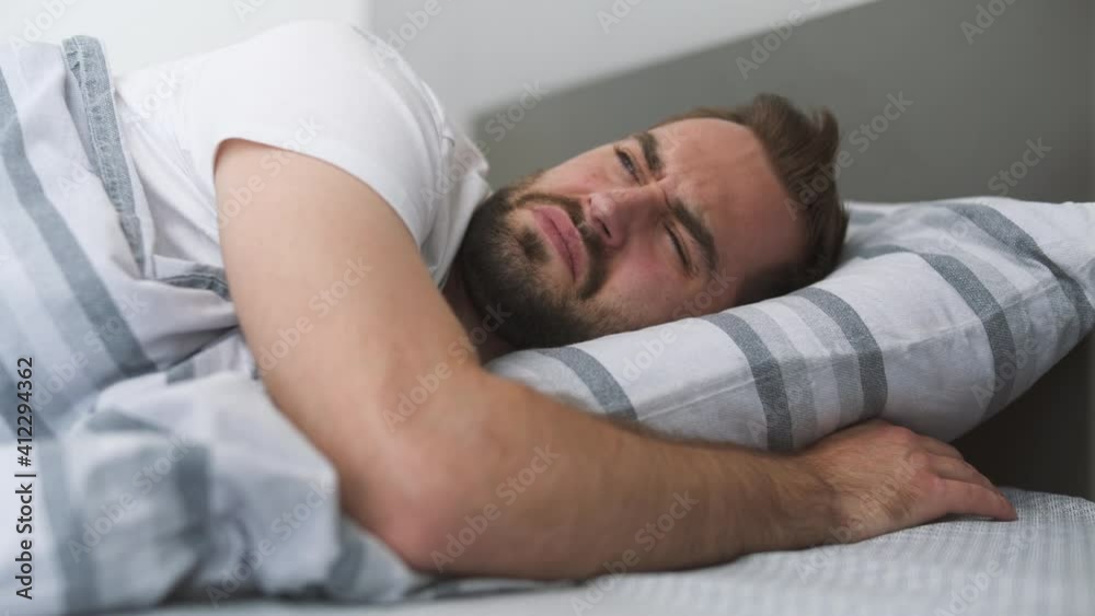 Tired irritated sad young man wear casual blue t-shirt lying in bed sleep with folded hands under pillow rest spend time in bedroom lounge home in own room house wake up open eyes. Real estate concept Tired irritated sad young man wear casual blue t-shirt lying in bed sleep with folded hands under pillow rest spend time in bedroom lounge home in own room house wake up open eyes. Real estate concept