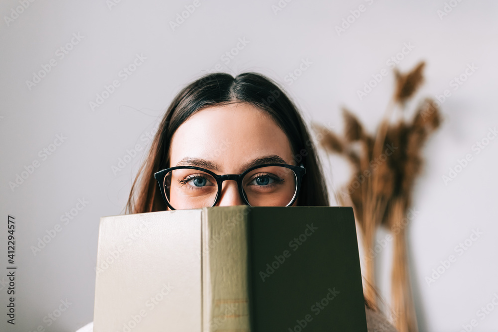 Portrait of young caucasian woman college student in eyeglasses hiding behind a book and looking at camera. - obrazy, fototapety, plakaty 