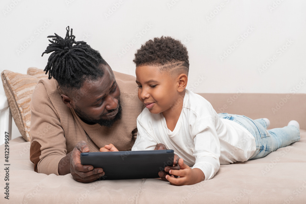 Fototapeta premium Dark skinned African male and his little son lie on the sofa at home and use tablet