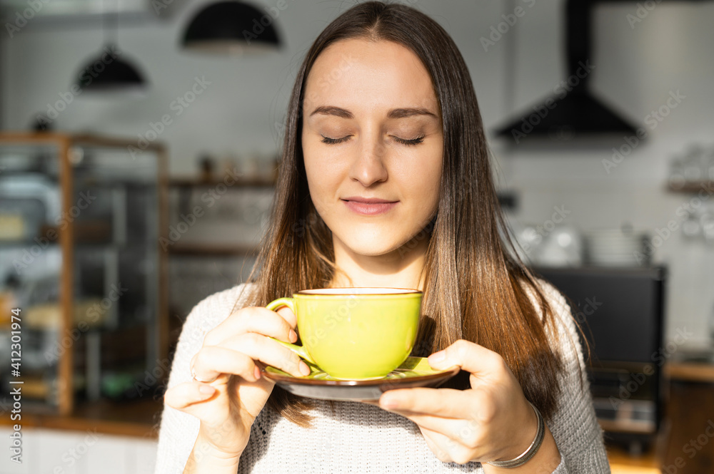 A young satisfied woman with eyes closed sniffs a fresh coffee from green mug sitting in the cozy cafe A young satisfied woman with eyes closed sniffs a fresh coffee from green mug sitting in the cozy cafe