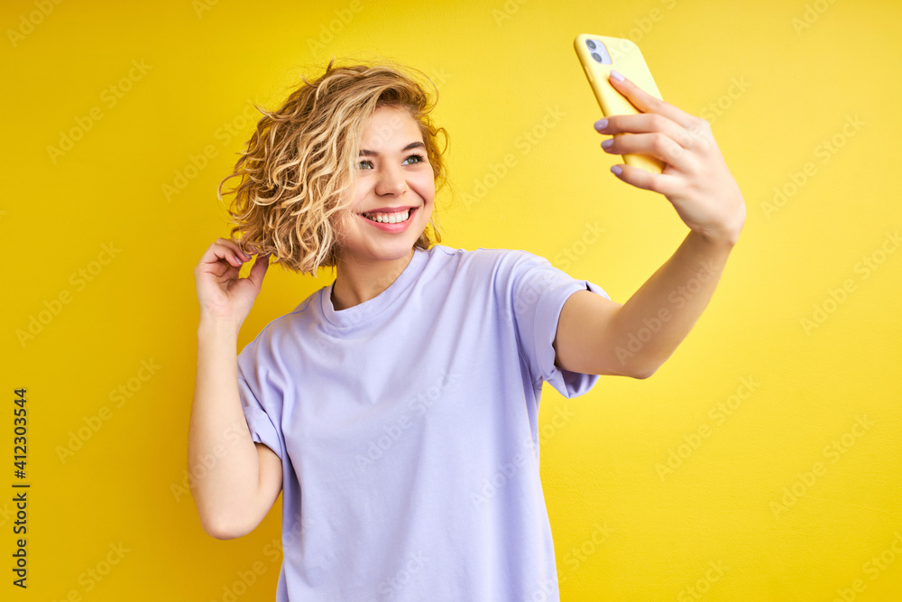 female posing making photo on smartphone, selfie, smiling. indoor studio shot isolated on yellow background