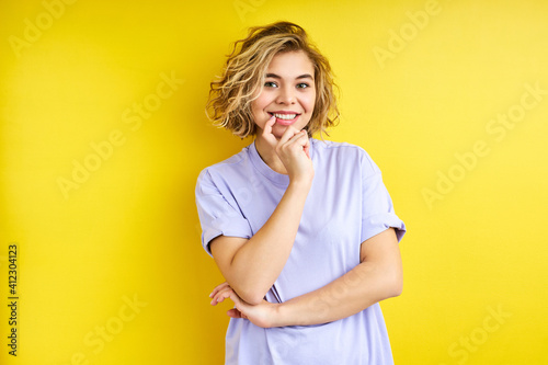teen of caucasian appearance posing isolated over yellow background, carefree woman holds finger on mouth and smile looking at camera. people concept