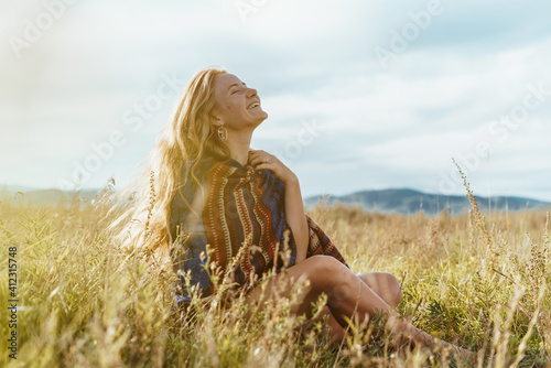 Natural natural beauty of the red-haired girl gleefully smiling sun and sky with an ethno cape on her shoulders sitting on the steppe grass against the background of hills, steppe and sky. High