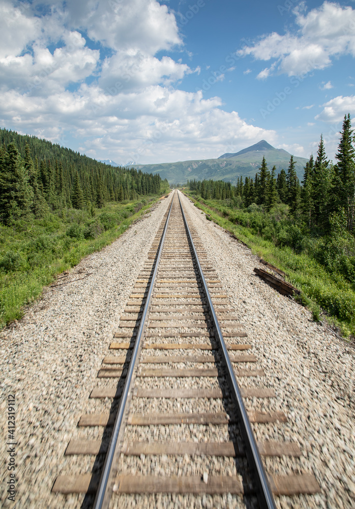 Railway tracks heading towards a mountain with clouds in the blue sky and trees beside the tracks