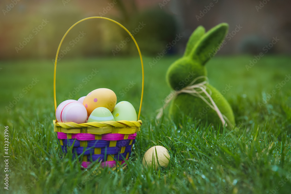 Easter basket with colored eggs and Easter bunny rabbit statuette on the green grass with dew. Easter egg hunt in the garden. Safe festive tradition during pandemic. Selective focus, copy space.