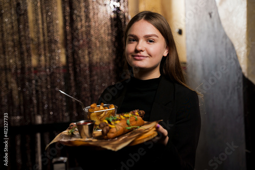 Waiter carries the order to the visitor. Barbecue grilled hot dog with yellow mustard and ketchup on wooden background. Hot dog sandwich with potato fries and sauces.