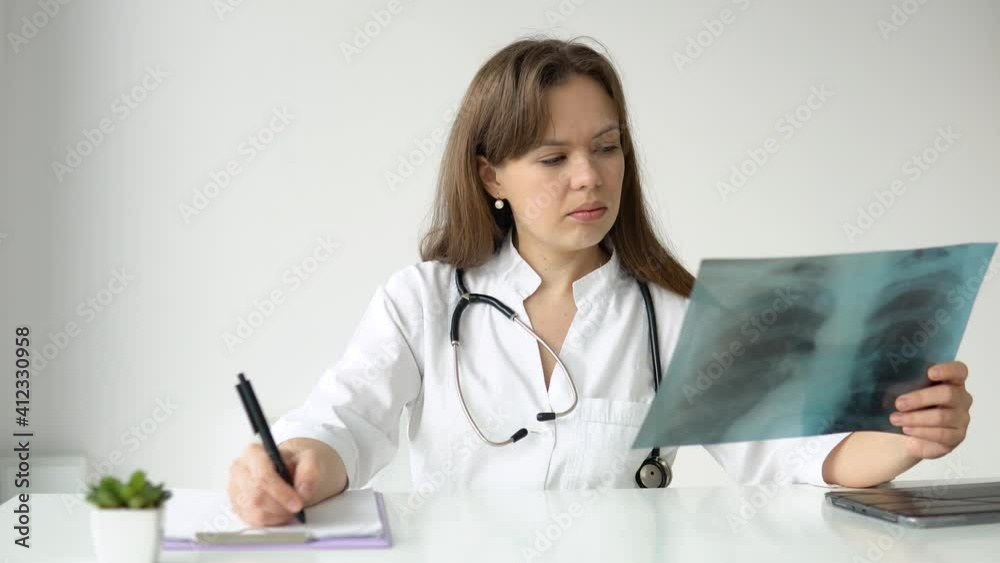 Young caucasian woman doctor wearing white medical coat and stethoscope writes a medical history in a notebook and holds the x-ray Young caucasian woman doctor wearing white medical coat and stethoscope writes a medical history in a notebook and holds the x-ray