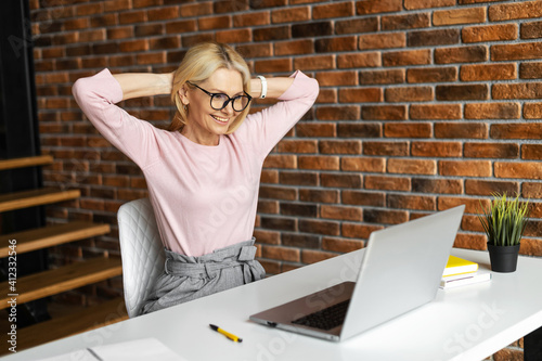 Mature mid-age businesswoman sitting at the desk, looking at the laptop and smiling, excited, satisfied and relaxed, enjoying the end of a workday, relaxed posture hands behind head. Well done concept photo