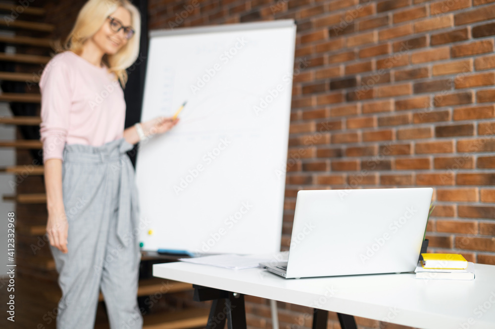 Mature female teacher, tutor coach in glasses with blond hair looking at the laptop screen, giving virtual lessons online by zoom conference call, or businesswoman presenting a project on a whiteboard Mature female teacher, tutor coach in glasses with blond hair looking at the laptop screen, giving virtual lessons online by zoom conference call, or businesswoman presenting a project on a whiteboard