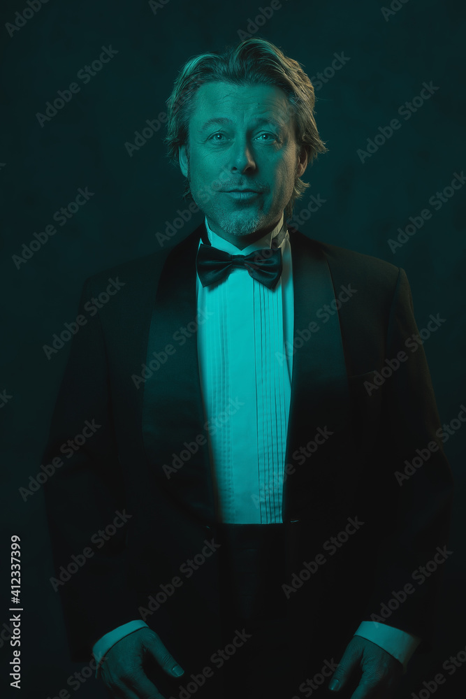 Fototapeta premium Blonde man wearing tuxedo and bow tie. Lit by blue and orange light against dark wall.