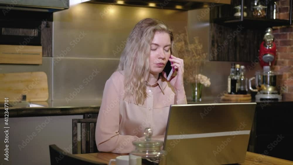 Portrait of girl working on a laptop in the evening at home in the kitchen. A female freelancer talks on a mobile phone and uses a computer to take notes. Discuss the project with the client remotely