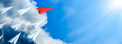 Red Paper Airplane Leading Fleet Of Small White Planes Away From Approaching Storm Toward Blue Sky And Sun - Leadership Concept
 photo