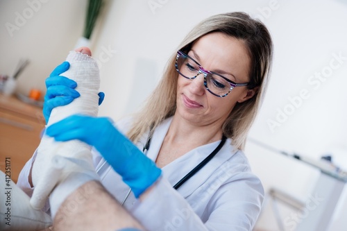 Smiling professional nurse with glasses is bandaging the leg. Fracture of human lower limbs.